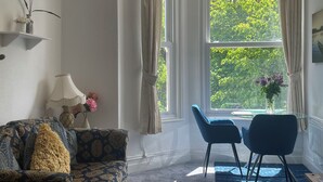 A room with a patterned sofa, a dining table with chairs, a vase with flowers, and a window with curtains. - Apartment 6 (Torquay)
