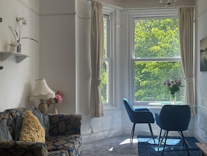 A room with a patterned sofa, a dining table with chairs, a vase with flowers, and a window with curtains. - Apartment 6 (Torquay)