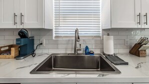 Een keuken met witte kastjes, een roestvrijstalen spoelbak, een koffiezetapparaat en een raam met jaloezieën.