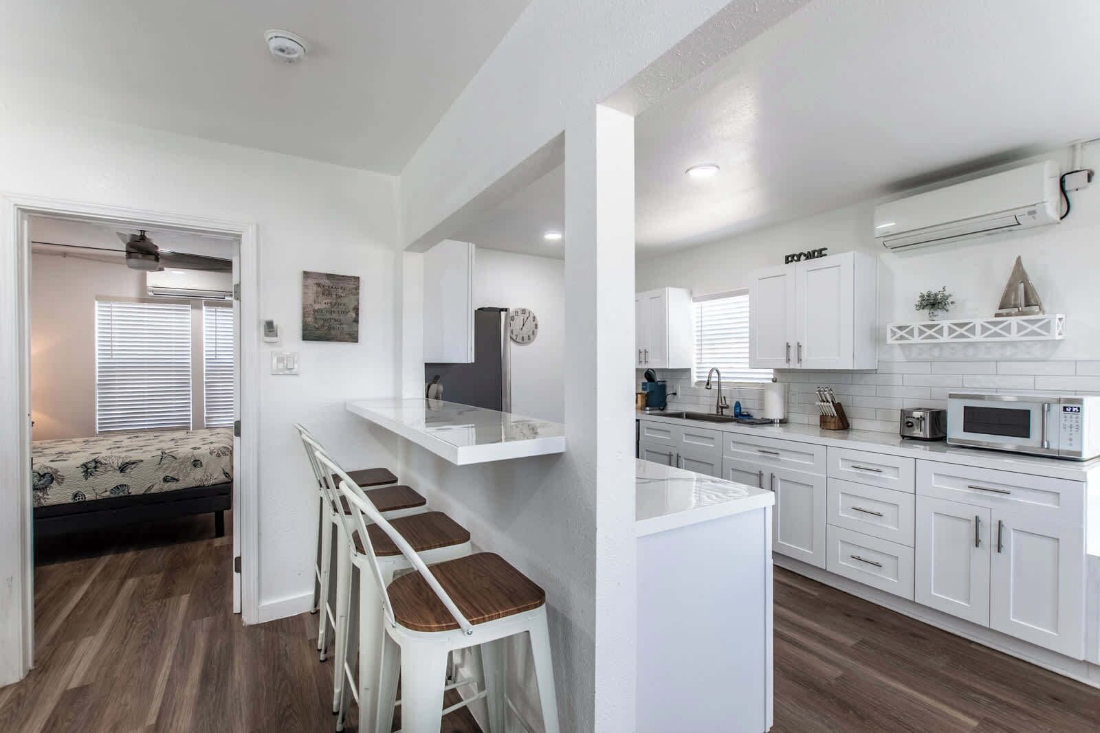 Una cocina moderna con armarios blancos, un microondas y un lavamanos. Se observa una pequeña isla con taburetes y un refrigerador.