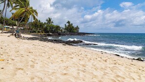 A sandy beach with palm trees, ocean waves, and a rocky shoreline. - Kona Reef#B2: Steps from Beach, Fully Remodeled, Ground Floor (Kailua Kona)