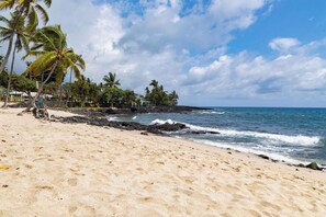Ein sandiger Strand mit Palmen, Wellen im Meer und einer felsigen Küstenlinie.