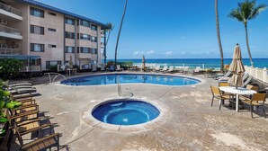 A pool area with lounge chairs and umbrellas, a building with balconies, and a view of the ocean. - Kona Reef#B2: Steps from Beach, Fully Remodeled, Ground Floor (Kailua Kona)