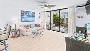A spacious living area with a sofa, coffee table, and chairs, a ceiling fan, a painting, and a view through a glass door to an outdoor seating area. - Kona Reef#B2: Steps from Beach, Fully Remodeled, Ground Floor (Kailua Kona)