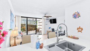 A modern kitchen with a stainless steel sink, a ceiling fan, and a dining area with a table and chairs. - Kona Reef#B2: Steps from Beach, Fully Remodeled, Ground Floor (Kailua Kona)