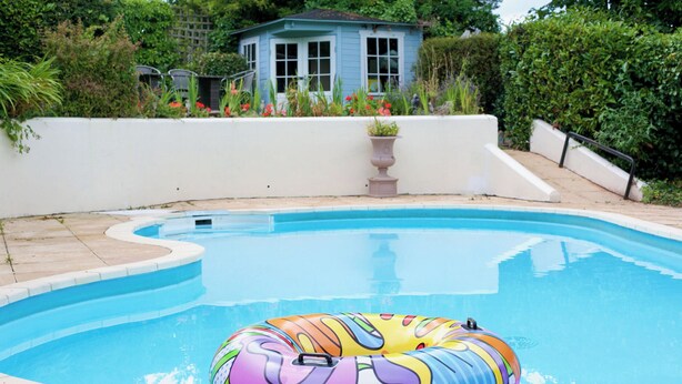 A swimming pool with a colorful inflatable ring in the water, surrounded by a white wall and a small blue house in the background. - Apartment 2 (Torquay)