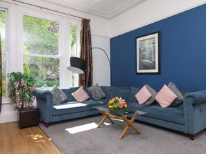 A living room with a blue sofa, a glass coffee table, a potted plant, and a painting on the wall. - Apartment 2 (Torquay)