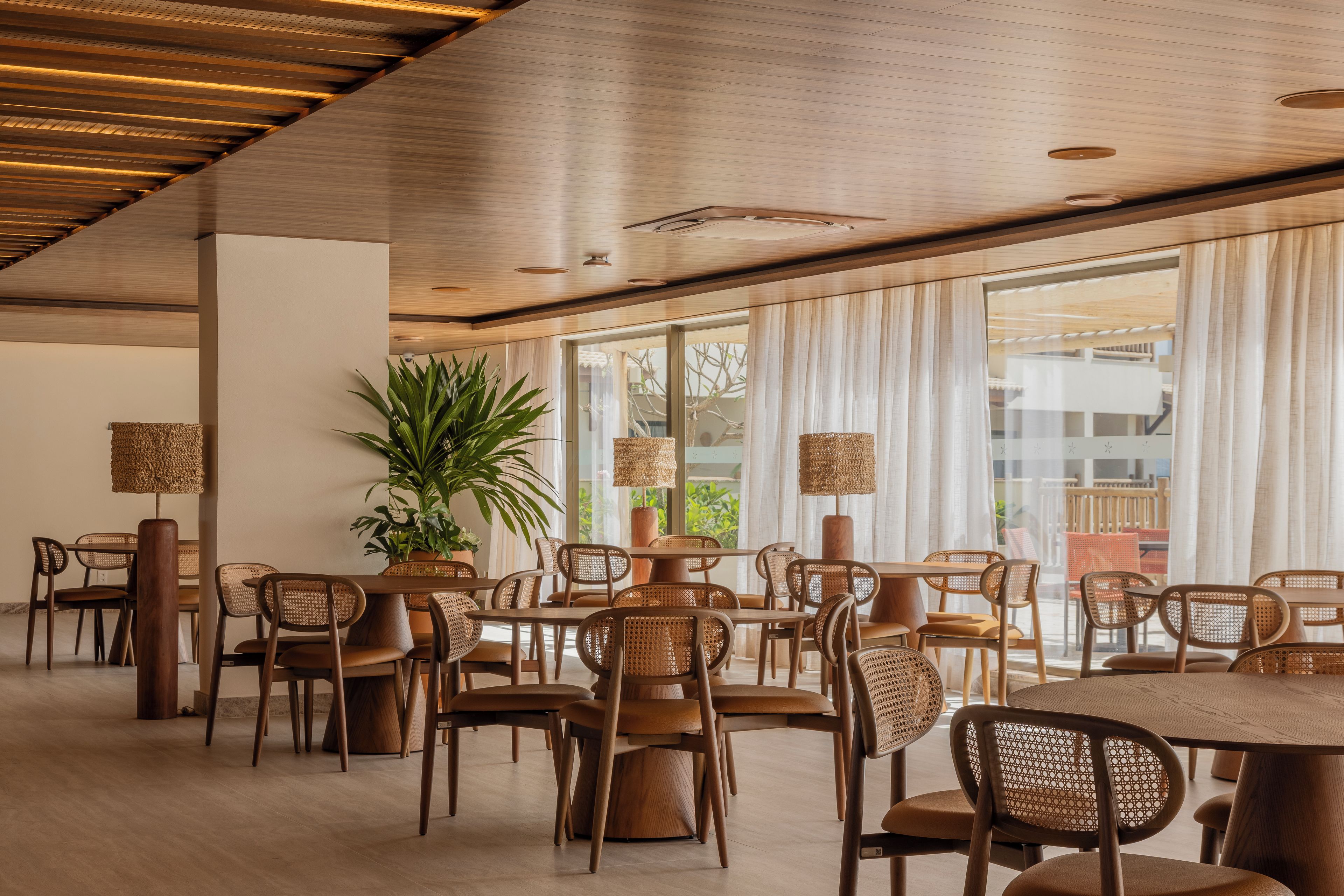 A spacious dining area with wooden tables and chairs, large windows with curtains, and a potted plant.