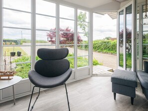 A room with large windows, a grey upholstered chair, a grey ottoman, and a small table with a ship model. - 4 Star Holiday Home in Haderslev (Haderslev)