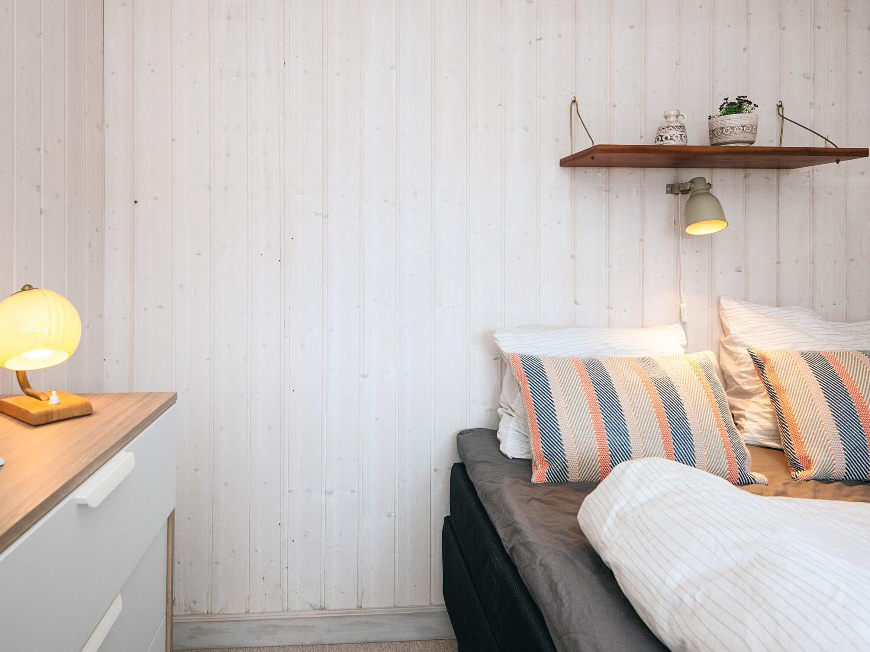 A bedroom with a bed, pillows, a bedside lamp, a wooden nightstand, and a shelf with decorative items.