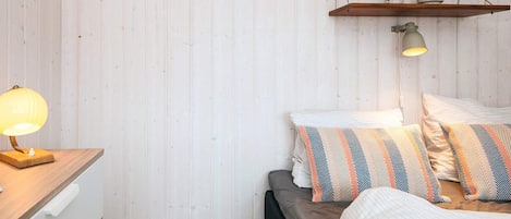 A bedroom with a bed, pillows, a bedside lamp, a wooden nightstand, and a shelf with decorative items.