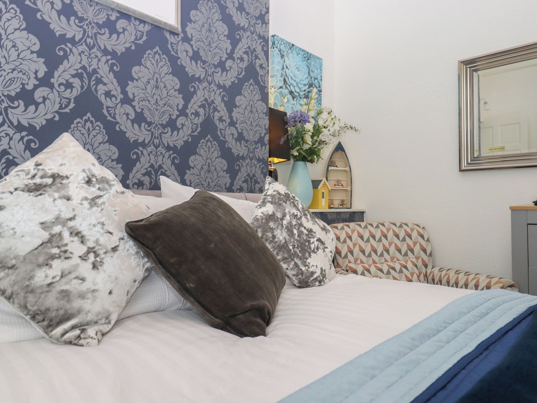 Une chambre à coucher avec un lit, des oreillers, un vase de fleurs, une petite étagère et un miroir.