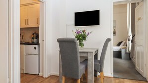 A compact living space with a kitchenette, a dining area with two chairs, a flat-screen TV mounted on the wall, and a small table with a vase of flowers. - Apartment 4 (Torquay)
