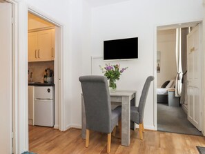 Ein kompakter Wohnraum mit Küchenzeile, einer Essecke mit zwei Stühlen, einem flachbildfernseher an der Wand und einem kleinen Tisch mit Blumenvase.