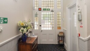 A hallway with a white door, a fire exit sign, a no smoking sign, a vase with flowers, and a small table with books. - Apartment 4 (Torquay)