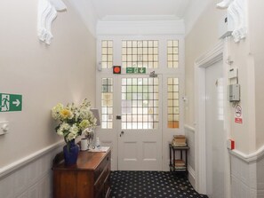 A hallway with a white door, a fire exit sign, a no smoking sign, a vase with flowers, and a small table with books. - Apartment 4 (Torquay)