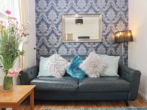 Ein dunkles Ledersofa mit flauschigen Kissen, ein gerahmter Spiegel, eine Blumenvase und eine Stehlampe.