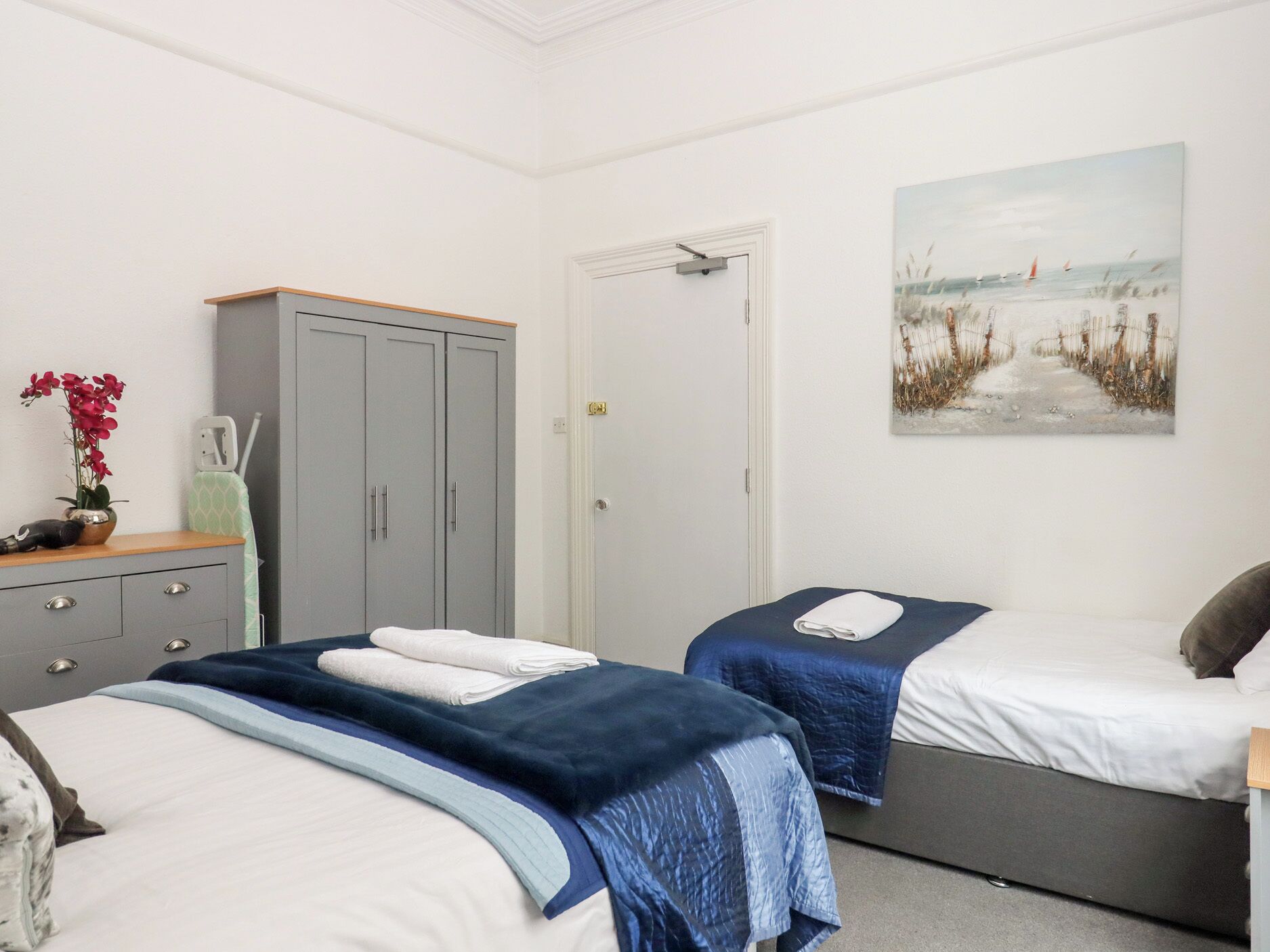 A bedroom with two beds, a wardrobe, a door, and a beach-themed painting.