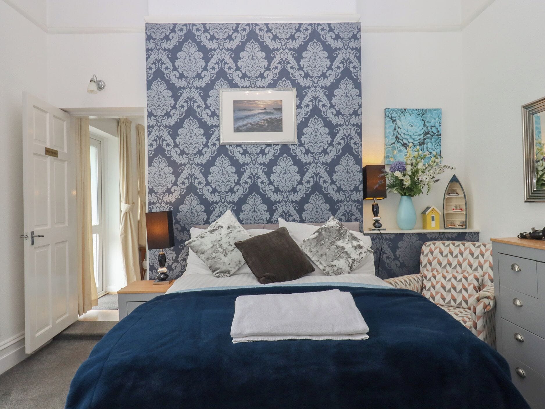Une chambre à coucher avec un papier peint à motifs, un lit recouvert d’une couverture bleue, une table de chevet avec une lampe, une commode et une porte donnant sur une autre pièce.