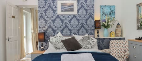 A bedroom with a patterned wallpaper, a bed with a blue cover, a nightstand with a lamp, a dresser, and a door leading to another room.