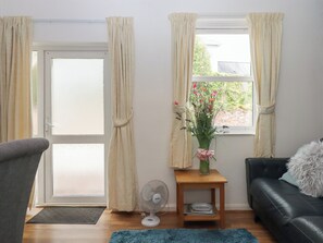 Ein Wohnzimmer mit einer Couch, einem kleinen Tisch mit Ventilator, einer Vase mit Blumen und einem Fenster mit Vorhängen.