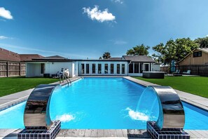 A rectangular swimming pool with a clear blue water, surrounded by a wooden deck and a white building with large windows. - Pearl Grand on Ocean (Corpus Christi)