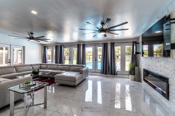 A spacious living room with a sectional sofa, a glass coffee table, a fireplace, and a pool visible through the glass doors. - Pearl Grand on Ocean (Corpus Christi)