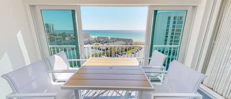 A balcony with a table and chairs, offering a view of the beach and buildings.