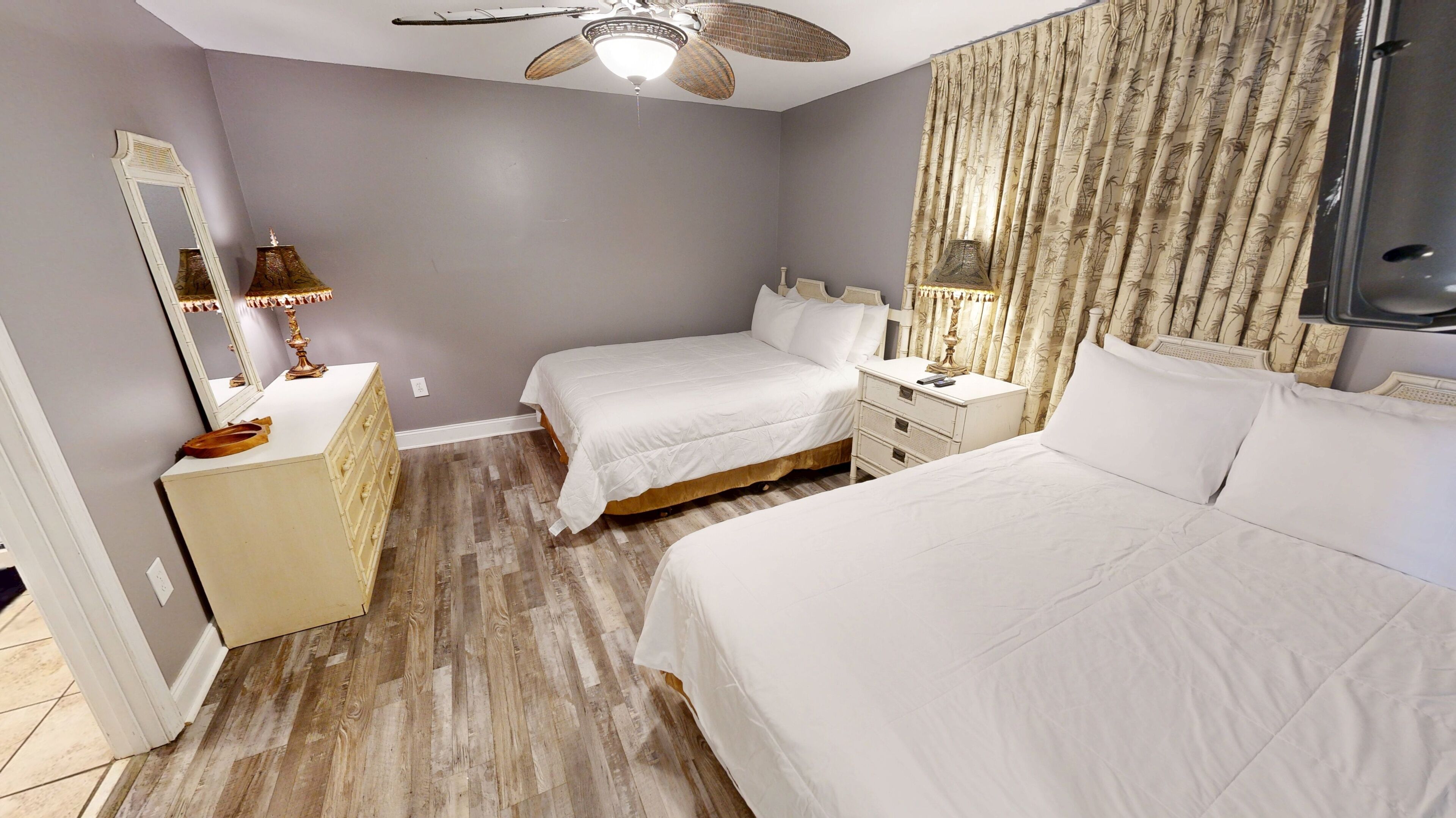A hotel room with two beds, a vanity with a mirror, a ceiling fan, and a large window with curtains.