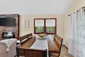 A dining area with a wooden table, chairs, a decorative bowl, a television on a brick wall, and a window with blinds.
