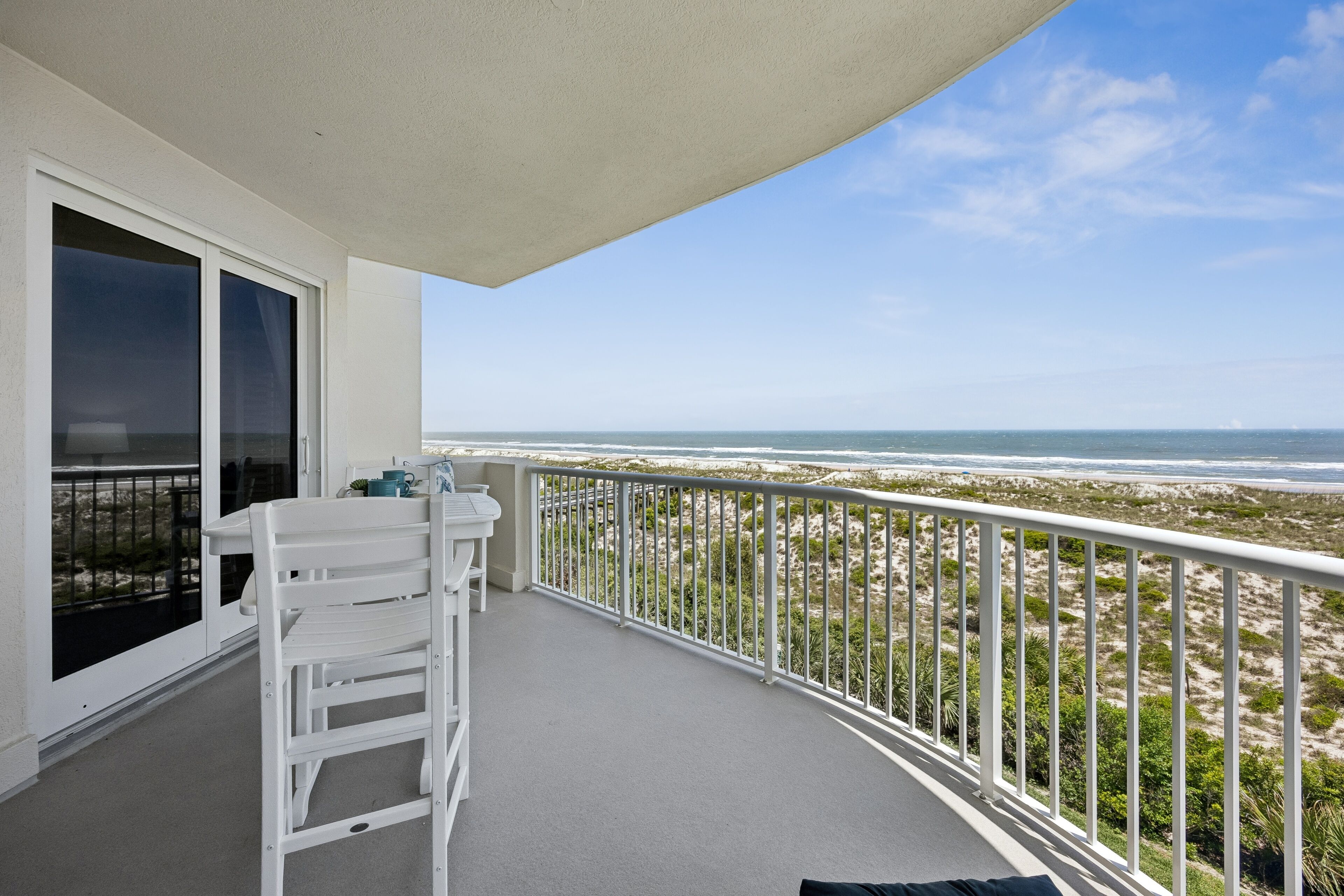 A balcony with a view of the beach, a white chair, and a table.