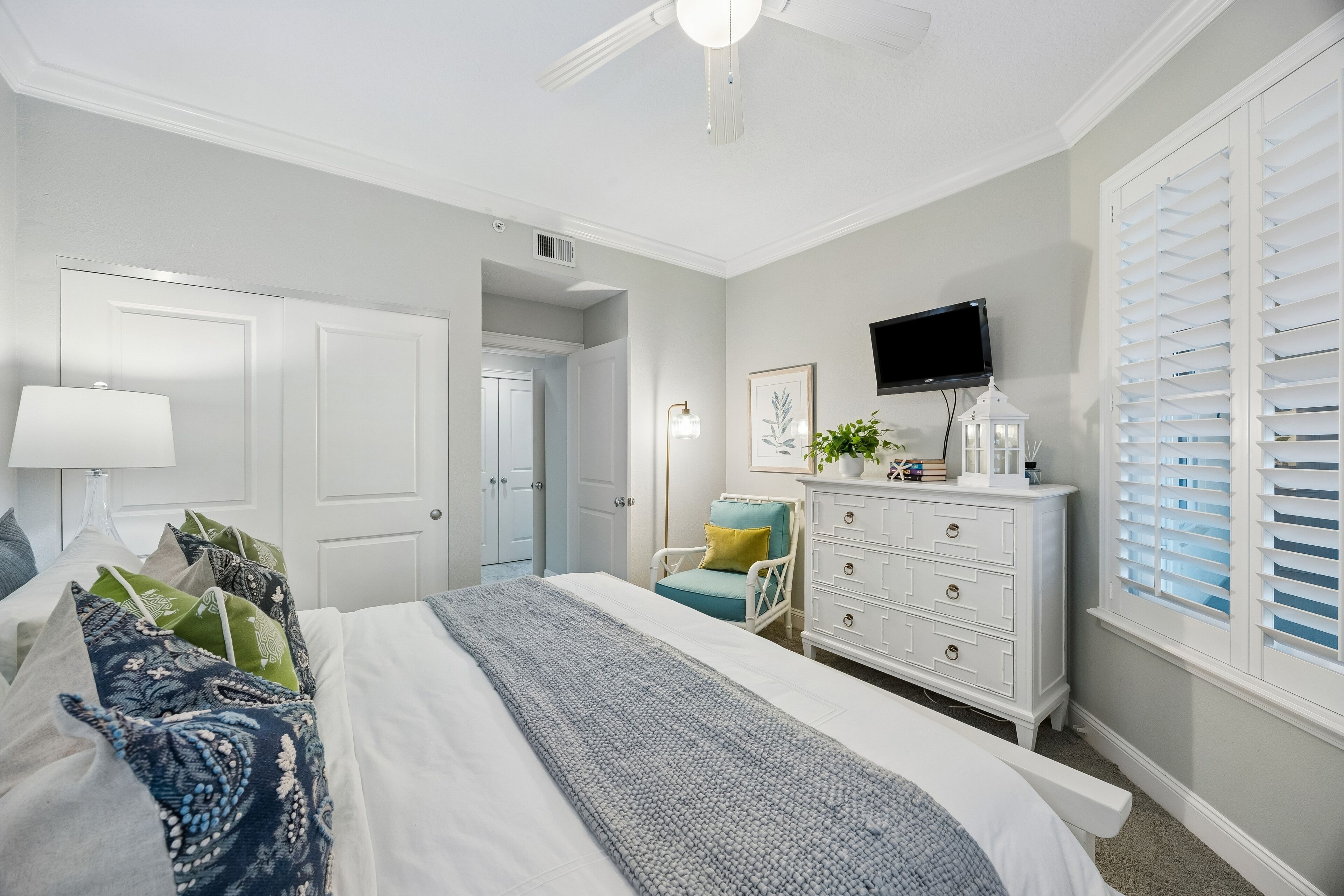 A bedroom with a bed, dresser, chair, TV, and window with blinds.
