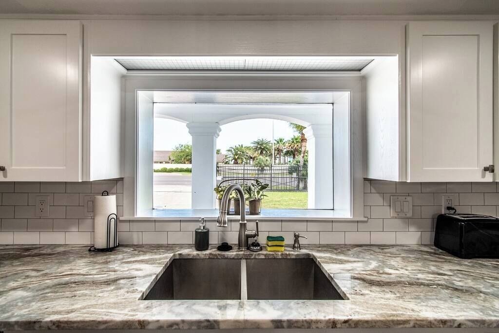 A kitchen with a large window, white cabinets, a double sink, and a marble countertop.