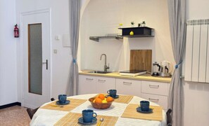 Eine kompakte Küche mit Spüle, kleiner Arbeitsfläche und einer Ablage darüber. Ein Tisch mit einer Schüssel Obst und blauen Tassen steht daneben.