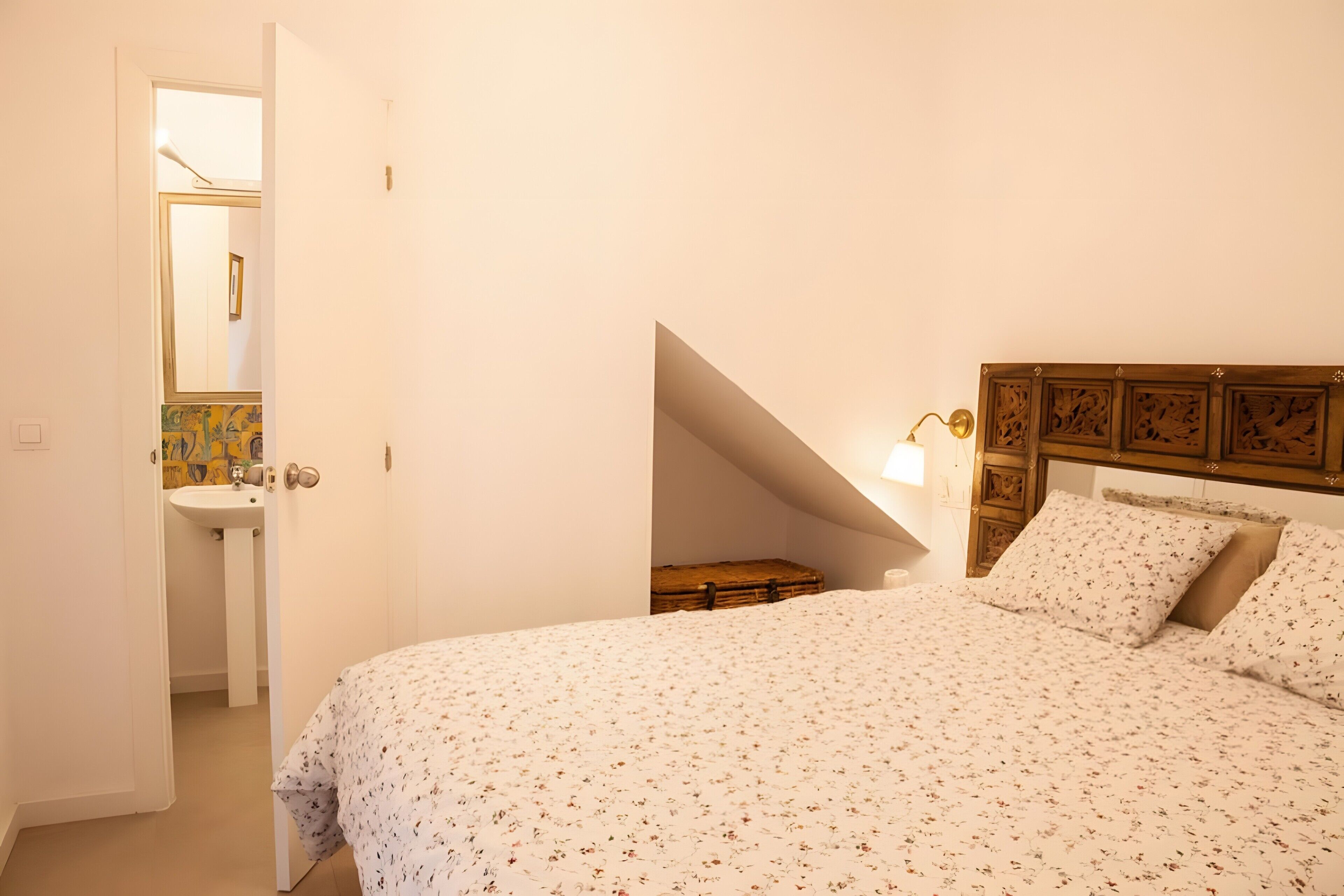 A bedroom with a bed, a nightstand, a wooden headboard, a wall-mounted light, and a door leading to a bathroom.