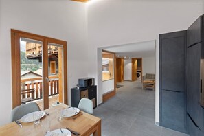 A modern kitchen with dark cabinetry, a dining area with a wooden table and chairs, and a view of a wooden deck and mountains in the background. - Charming apartment in an authentic village (Anniviers)