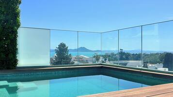 A clear view of a swimming pool with a wooden deck and a glass barrier, overlooking a scenic landscape with mountains and buildings.