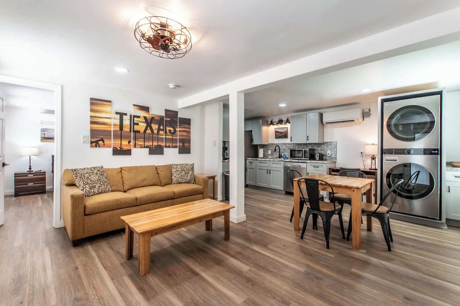 A modern living room with a sofa, coffee table, and dining area. A kitchen with appliances and cabinets is visible in the background.