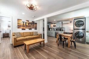 A modern living room with a sofa, coffee table, and dining area, connected to a kitchen with appliances and a laundry machine. - Serenity Suites C (Corpus Christi)