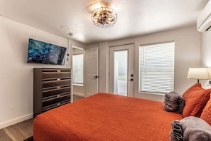 A bedroom with a large bed, a TV mounted on the wall, a ceiling light fixture, and a door with a window. - Serenity Suites C (Corpus Christi)