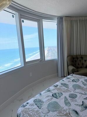 A bedroom with a large window overlooking a beach, a bed with a nautical-themed bedspread, and a tufted armchair. - Blue Horizon 2/2 Oceanfront AMAZING views, Pool, Hot tub, 27SE (Daytona Beach)