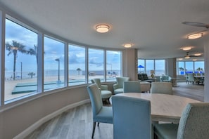 Ein geräumiges Zimmer mit großen Fenstern und Blick auf Strand und Palmen. Ein runder Tisch mit Stühlen sowie ein Deckenventilator sind vorhanden.