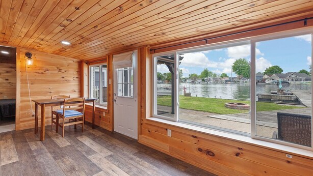 Une cabane aux murs et au plancher en bois, un coin repas avec une table et des chaises, et de grandes fenêtres offrant une vue sur un lac et un quai.