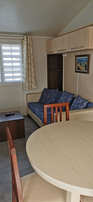 Ein Zimmer mit einem runden Holztisch, einer Couch mit gemusterten Kissen, einem Holzstuhl, einem kleinen Tisch mit Fernbedienung und einem Fenster mit Vorhängen.