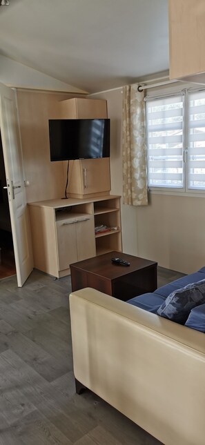 Ein Zimmer mit einer Couch, einem Fernseher auf einem Schrank, einem Fenster mit Vorhängen und einer Tür.