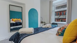 A bedroom with a bed, a large mirror, a window with a view of buildings, and a blue accent wall. - Spacious 2BR Family Home in Brierfield Near Burnley and Skipton (Burnley)