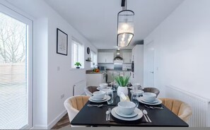 Un coin repas avec une table dressée pour quatre personnes, un luminaire suspendu moderne et une vue sur la cuisine adjacente.