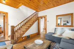 Una escalera de madera que lleva a un piso superior, un sofá gris con una manta de piel y una mesa de centro de madera con un plato decorativo.