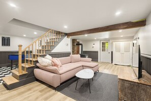 A modern living room with a wooden staircase, a pink sectional sofa, a small white table, and a flat-screen TV. - Heritage home with spa, EV charger and walkable village (Potton)