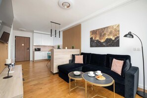 A modern living area with a dark sofa, a small round table with a cup and saucer, and a kitchen in the background. - Luxury Studio with Parking | Baltic Waves Resort | Near the Beach (Międzyzdroje)